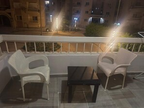 A balcony with two white plastic chairs and a small black table, overlooking a street with buildings and streetlights. - Modern 2BR in 6th October Balcony, Parking, WiFi & Smart TV (6th October)