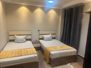 A hotel room with two single beds, a nightstand, a small table, and a dark curtain. - Modern 2BR in 6th October Balcony, Parking, WiFi & Smart TV (6th October)
