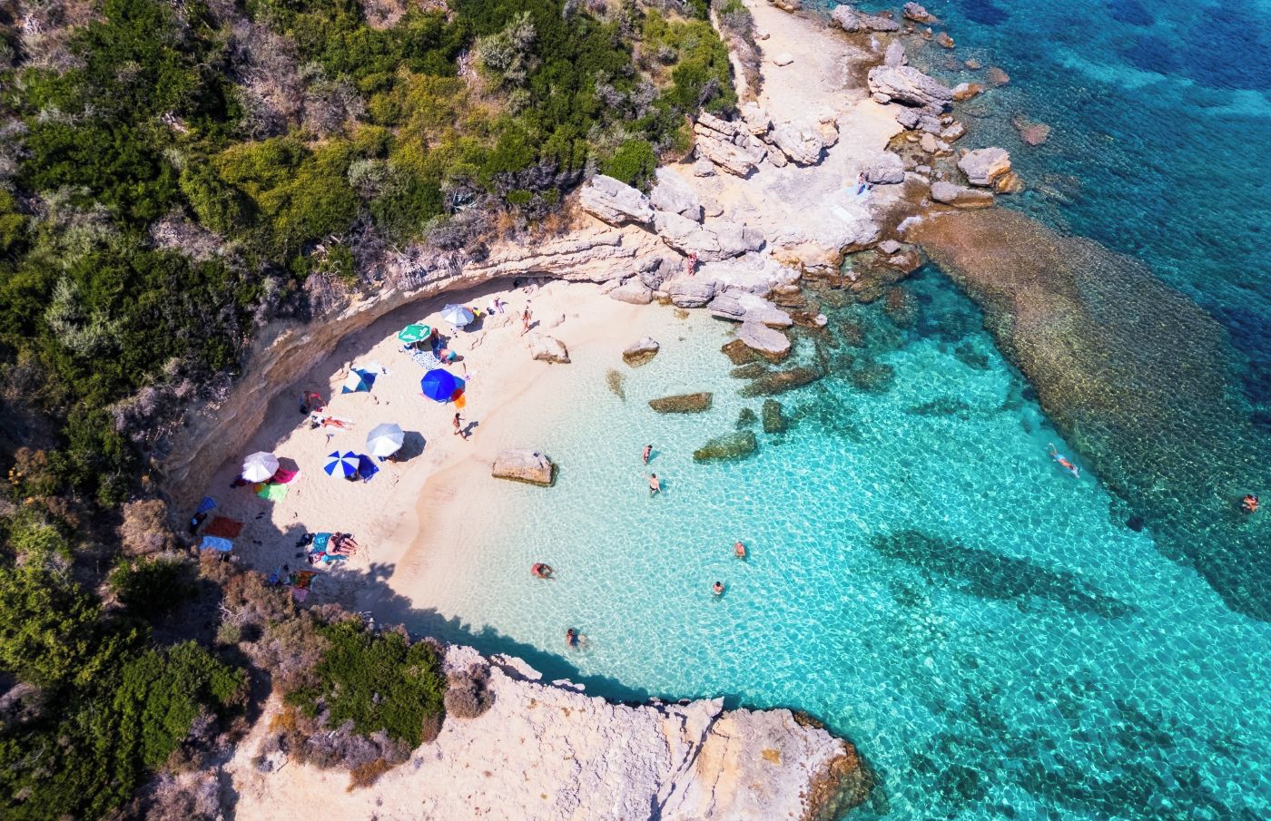An aerial view of a beach with clear turquoise waters, rocky formations, and a sandy shore with umbrellas and people.