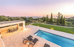 Un área de piscina moderna al aire libre con zona de comedor y sillones de descanso, con vistas a un paisaje pintoresco con árboles y el mar.