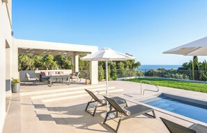 A modern outdoor pool area with lounge chairs and a covered seating area, overlooking a landscaped garden and the sea. - Stylish New Villa with Sea Views Walk to Beach (Livathou)