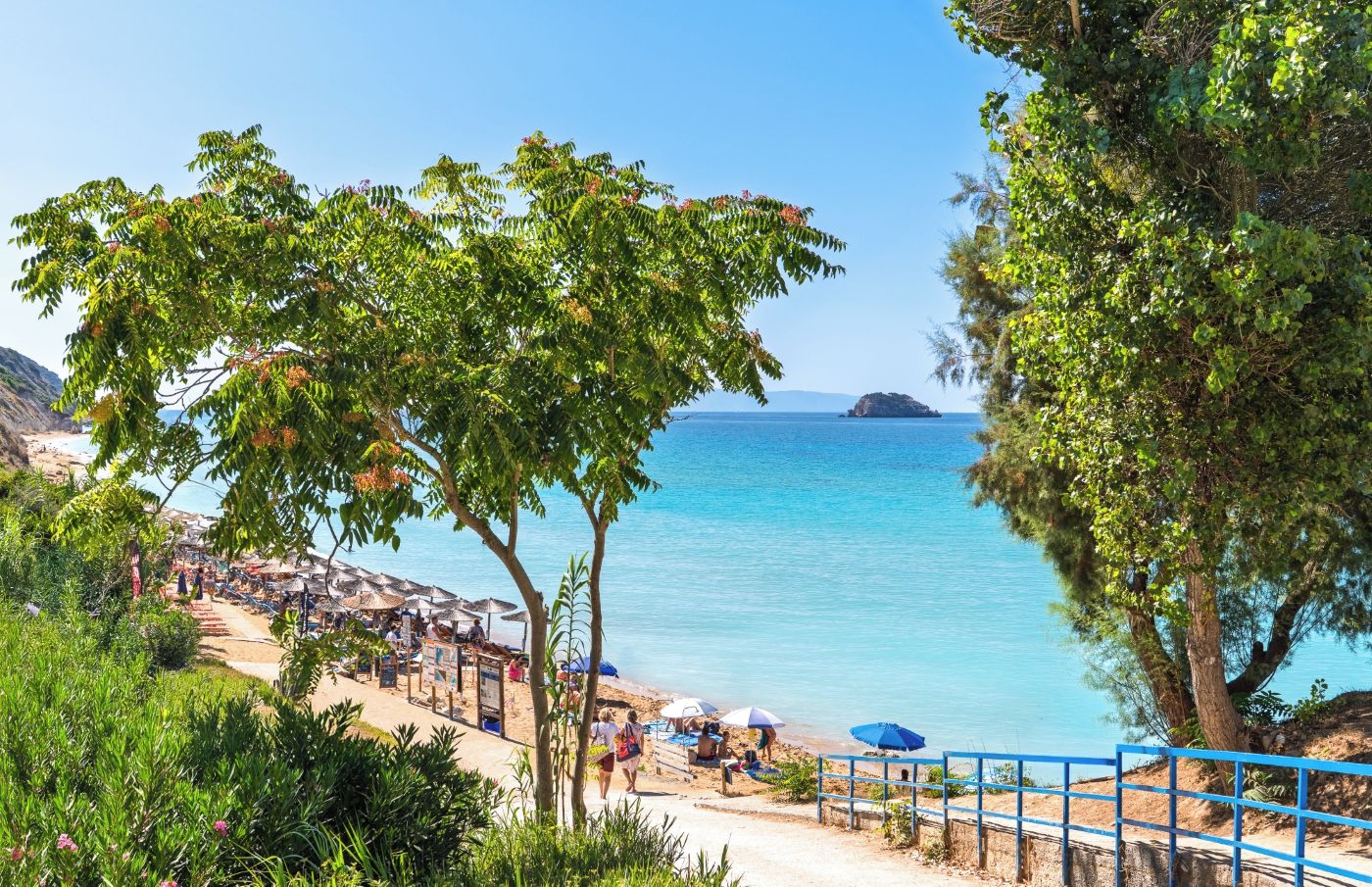 Una playa con vista despejada al mar, una pequeña isla a lo lejos y algunas personas relajándose bajo sombrillas.