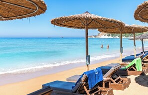 Une plage avec des chaises longues et des parasols en chaume, une mer d’un bleu limpide et, au loin, des formations rocheuses.