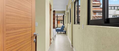 A corridor with a wooden door, a bed with a blue cover, and a window with a view of buildings.