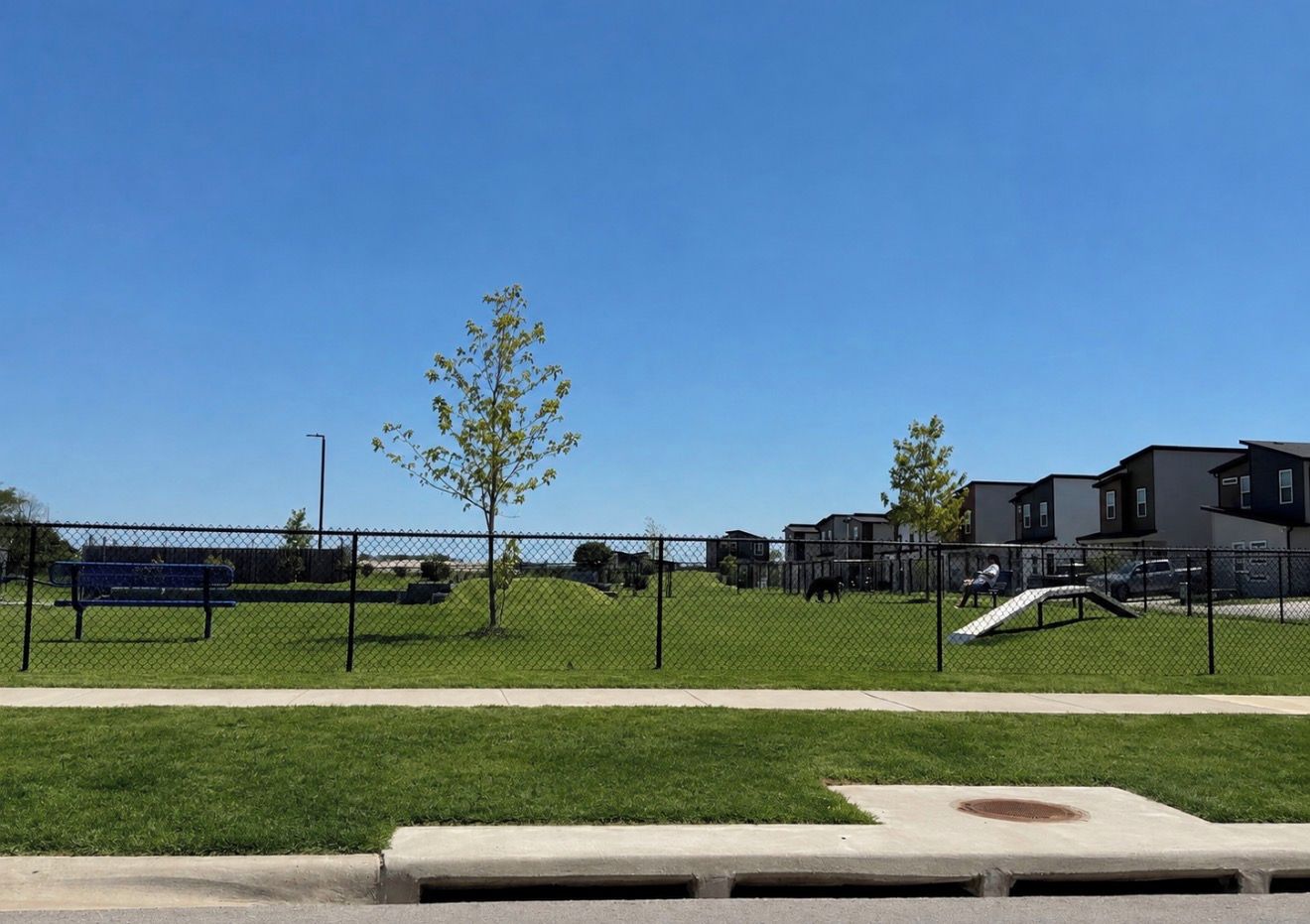 Ein von einer Hecke umgebener Park mit Spielplatz, Bänken und blauem Himmel.