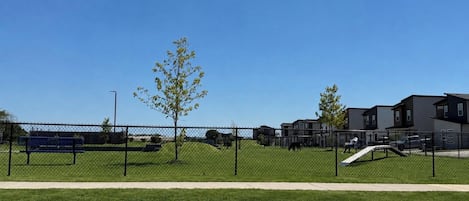 Ein von einer Hecke umgebener Park mit Spielplatz, Bänken und blauem Himmel.
