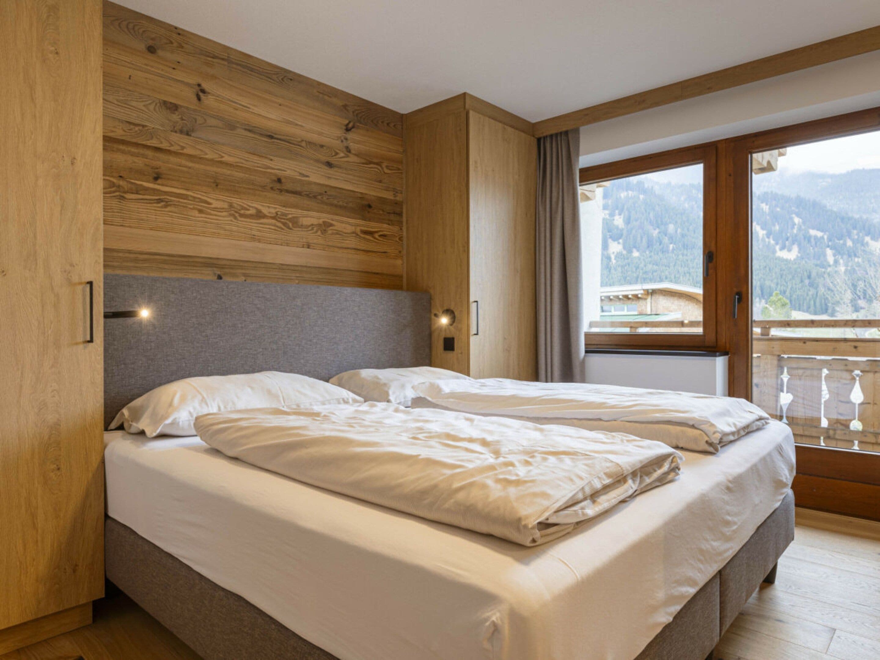 A double bed with a grey headboard in a room with wooden paneling and a view of mountains through a large window.