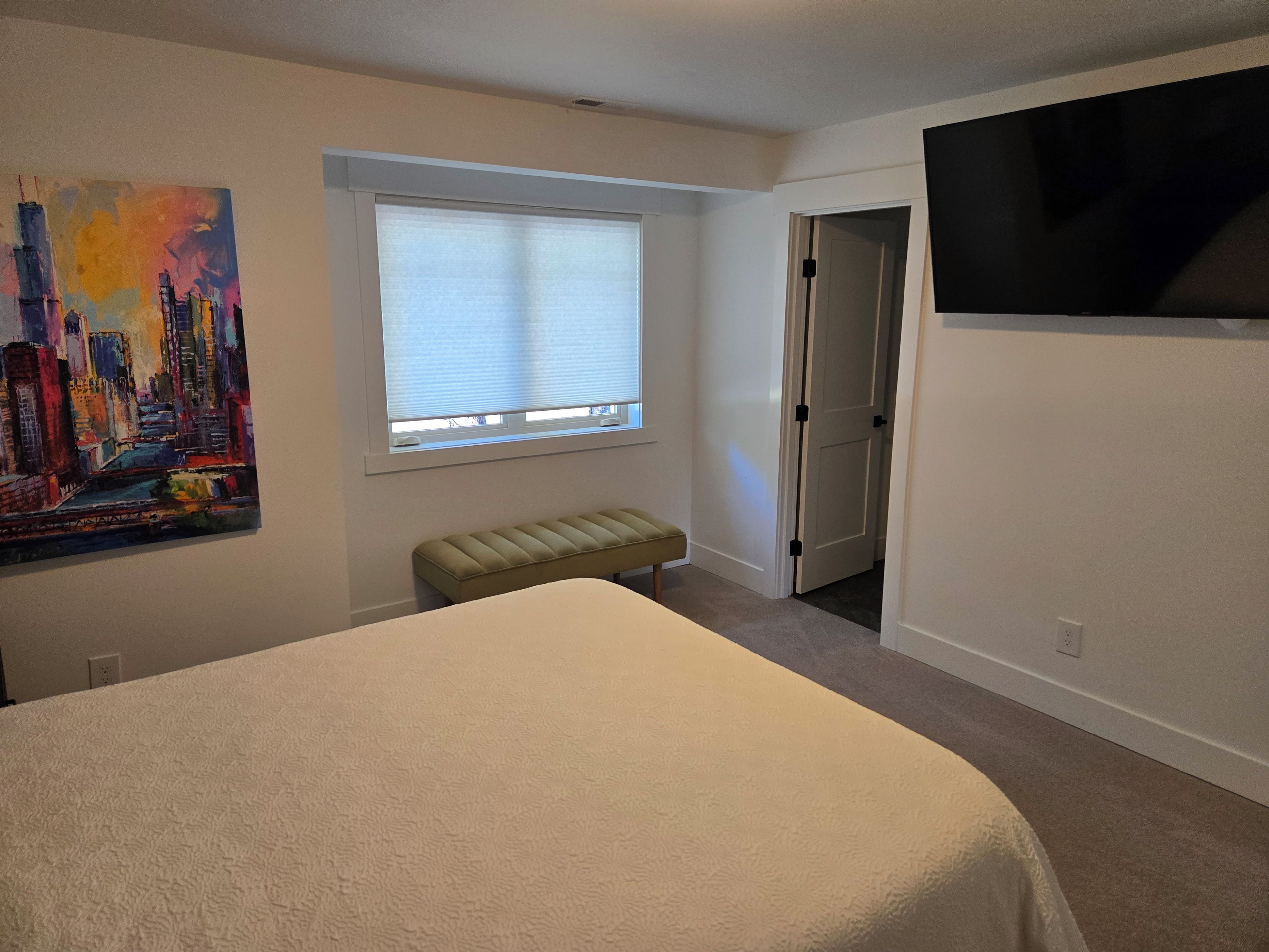 A bedroom with a bed, a window with blinds, a bench, a door, and a painting on the wall.