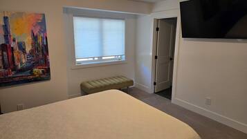 A bedroom with a bed, a window with blinds, a bench, a door, and a painting on the wall.