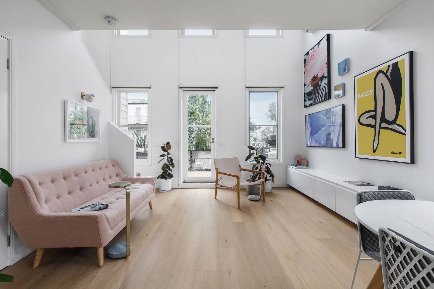 Una sala de estar moderna con un sofá rosa, una silla de madera y una mesa de comedor. Hay ventanas amplias y obras de arte en las paredes.
