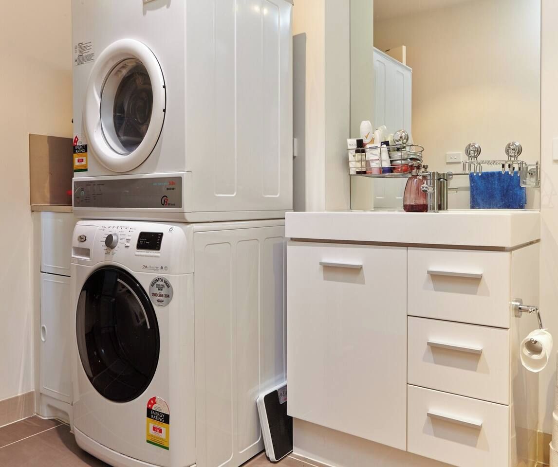 A compact laundry room with stacked washing machines, a small trash bin, and a white cabinet with drawers.