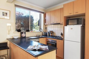 A kitchen with wooden cabinets, a black countertop, a white refrigerator, and a window with a view of trees. - Northcote Charm Spacious and Quiet Suburban Stay (Northcote)