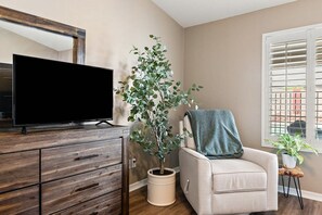 Un salon avec un téléviseur à écran plat posé sur un meuble en bois, une plante en pot, un fauteuil beige agrémenté d’un plaid et une fenêtre avec des stores.