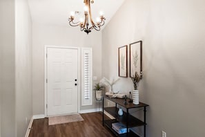 A hallway with a white door, a black console table with decorative items, and a chandelier. - Surprise House 🌵⛳ Fenced Pool & Firepit Near Surprise Stadium (Surprise)