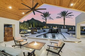 A modern outdoor patio with a fire pit, seating, and a view of a mountain at dusk. - Dos Palms | Luxury | HtdPool, Jacuzzi, Trails! (Phoenix)