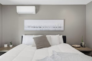 A modern bedroom with a large bed, a wall-mounted air conditioner, a framed abstract painting, and a bedside table with a plant and decorative items. - Dos Palms | Luxury | HtdPool, Jacuzzi, Trails! (Phoenix)
