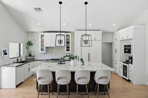 A modern kitchen with a central island, bar stools, pendant lighting, and white cabinetry. - Dos Palms | Luxury | HtdPool, Jacuzzi, Trails! (Phoenix)