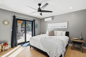 A modern bedroom with a bed, a ceiling fan, a window with curtains, and a view of the outdoors. - Dos Palms | Luxury | HtdPool, Jacuzzi, Trails! (Phoenix)