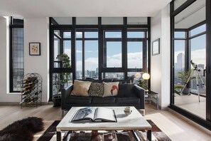 A modern living room with a black sofa, a coffee table with an open book, a wine rack, and large windows offering a city view. - Scenic Penthouse Living in the Heart of Melbourne (Southbank)