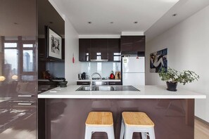 Une cuisine moderne avec des meubles de couleur sombre, un évier en acier inoxydable et un plan de travail blanc. On y trouve deux tabourets en bois et une plante en pot posée sur le plan de travail.
