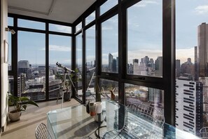 Ein Hotelzimmer in einem Hochhaus mit großen Fenstern und Blick auf die Stadt, ein Teleskop und ein Glastisch mit Stühlen.
