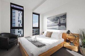 Ein modernes Schlafzimmer mit einem großen Bett, einem schwarzen Ledersessel, einem Nachttisch aus Holz mit einer Lampe und einem großen Fenster mit Blick auf die Stadt.