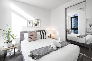 Une chambre d’hôtel moderne avec un grand lit, une table de chevet avec une lampe, une plante en pot et des œuvres d’art accrochées aux murs.