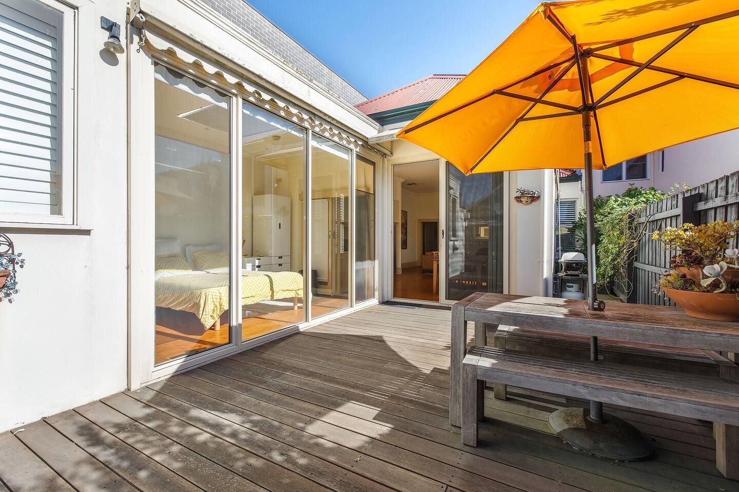 Eine Holzterrasse mit Tisch und Stühlen, einem gelben Sonnenschirm und einer Glastür, die zu einem Schlafzimmer mit Bett und Kommode führt.