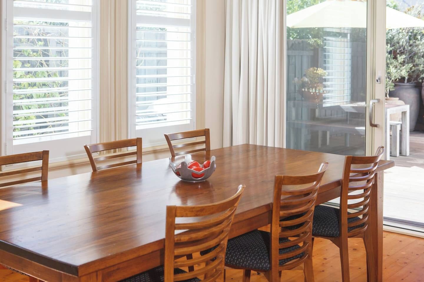 A dining area with a wooden table and six chairs, a bowl with red items, and a view of greenery through the window.