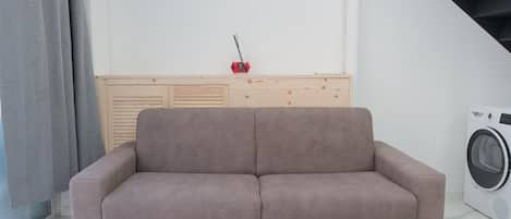 Een sofa in een neutrale tint in een moderne kamer, met een wasmachine en een houten kast op de achtergrond.