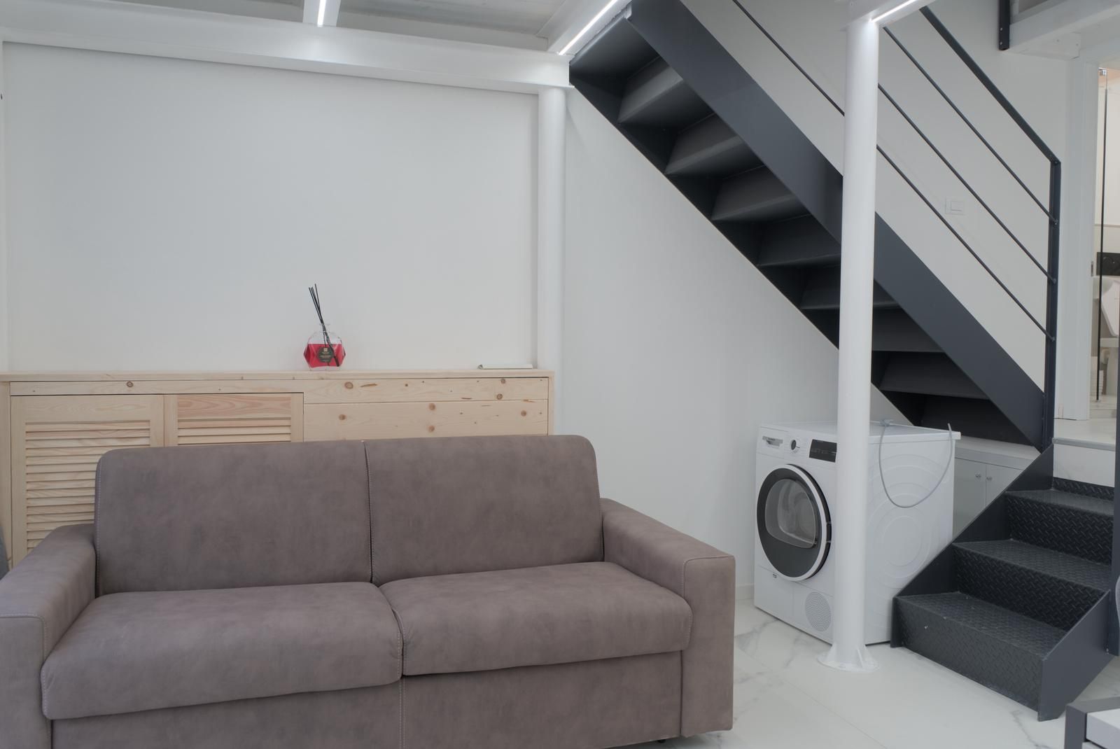Un espacio habitable moderno con un sofá gris, una lavadora y una escalera.
