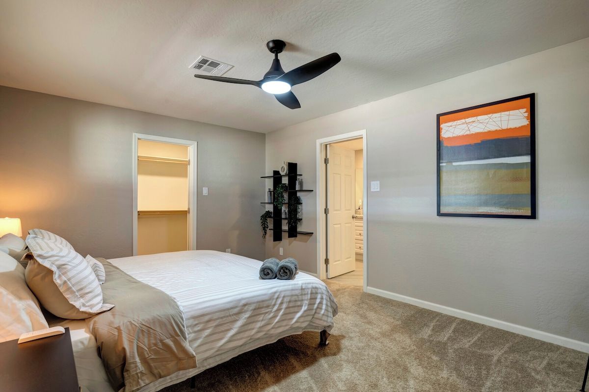 Une chambre à coucher avec un lit, un ventilateur de plafond, une table de chevet, une lampe, une porte, une étagère avec des plantes et un tableau au mur.