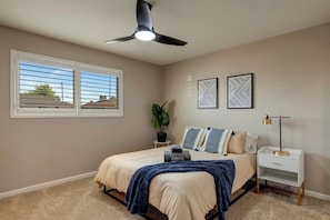 Une chambre à coucher comprenant un lit, des tables de chevet, un ventilateur de plafond, une fenêtre avec des stores et deux cadres photo fixés au mur.