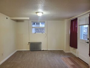 A room with a radiator, a window with blinds, a door with a curtain, and a ceiling light. - Fully Furnished 1-Bed Apartment in Cliff Cannon (Spokane)