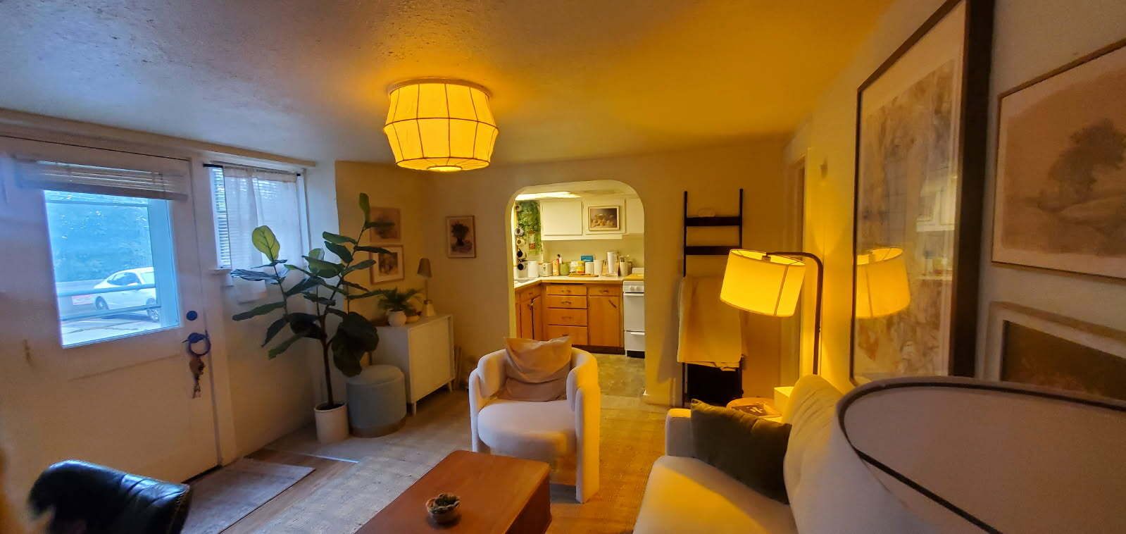 Una sala de estar acogedora con un sofá, una mesa de centro y una lámpara. Se ve una cocina al fondo.