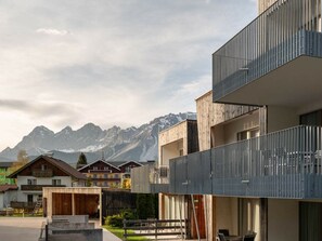 Un edificio moderno con balcón, una estructura de madera en primer plano y una cordillera al fondo.