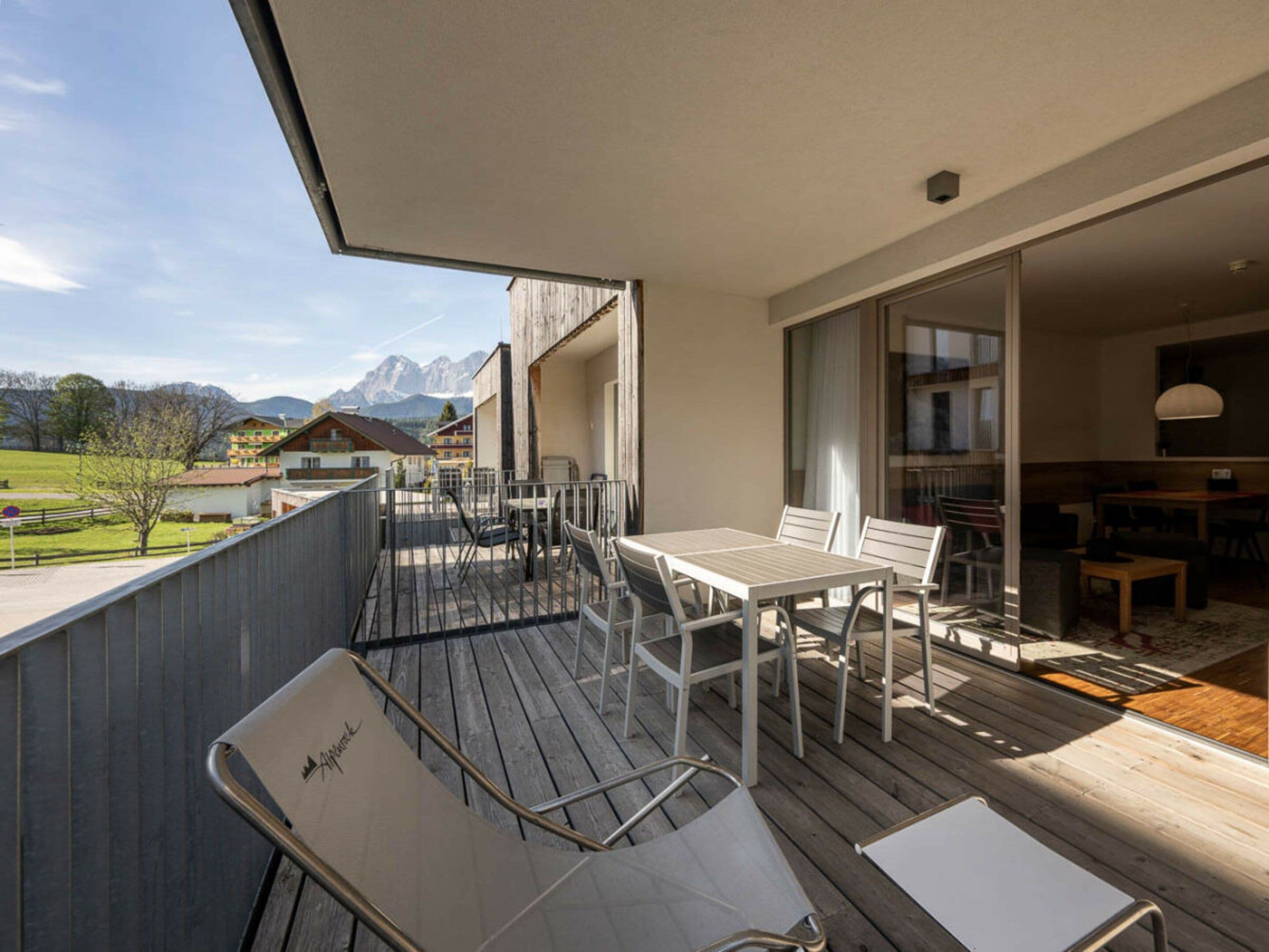 A balcony with a table and chairs, a view of the mountains, and a building with a wooden facade.