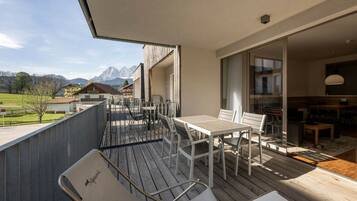 A balcony with a table and chairs, a view of the mountains, and a building with a wooden facade.
