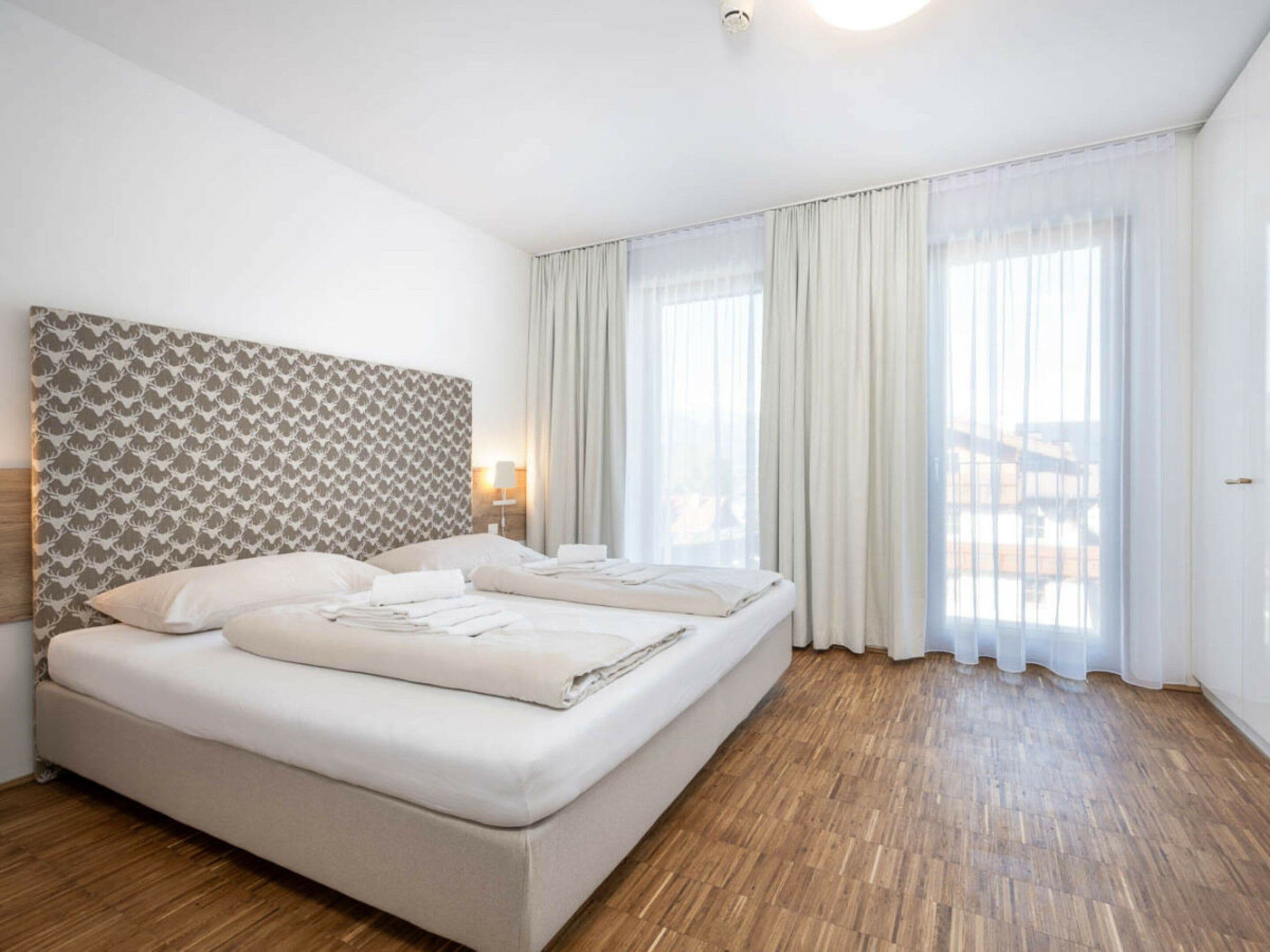 Ein Hotelzimmer mit einem großen Bett, einem gemusterten Kopfteil, einem Fenster mit leichtem Vorhang und einem Holzboden.