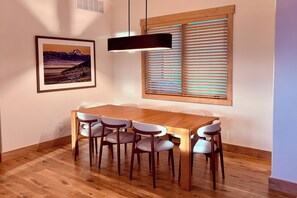 A dining area with a wooden table and chairs, a pendant light, a window with blinds, and a framed landscape painting on the wall. - The Oar Lock Cottage (Ennis)