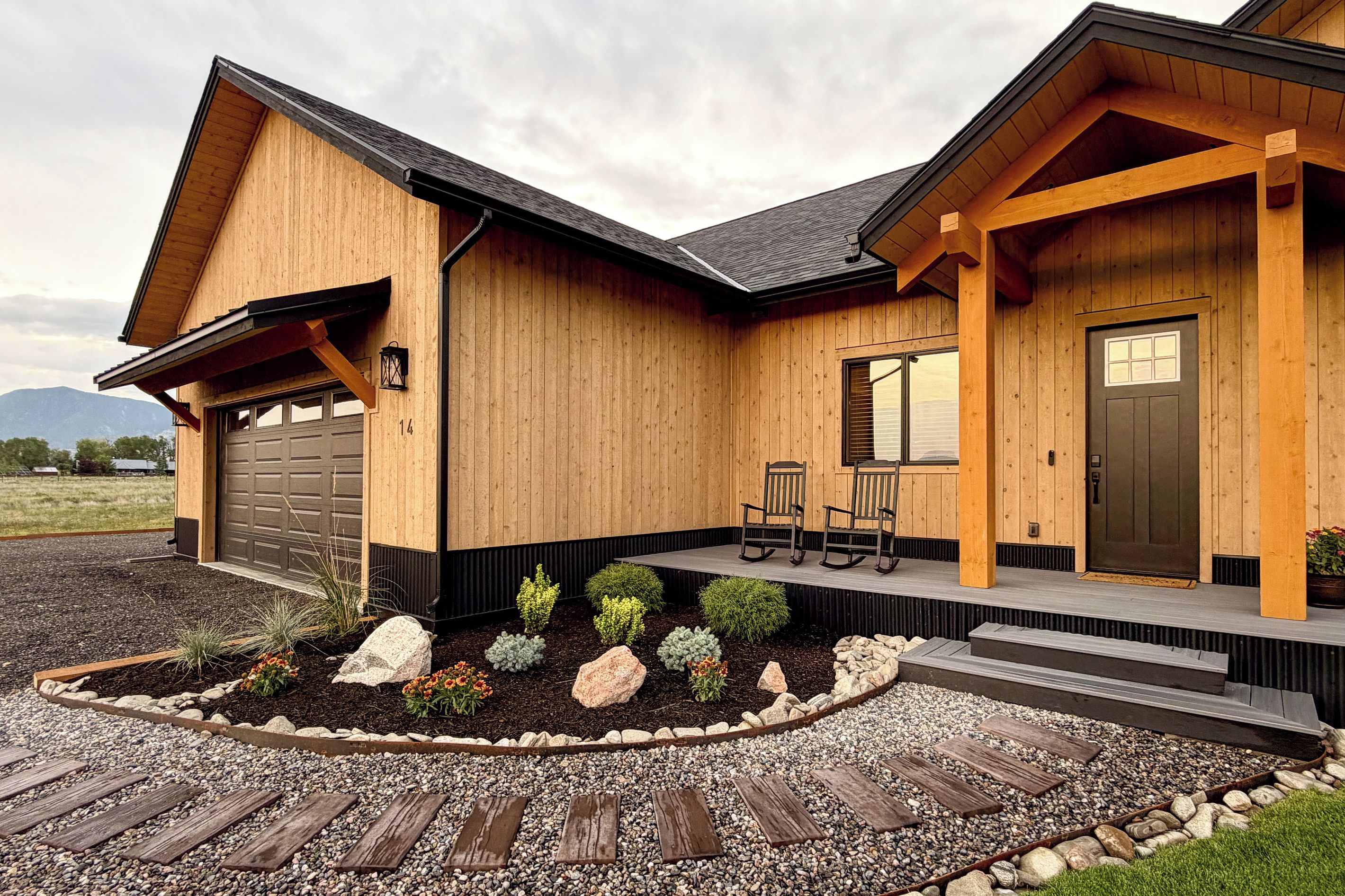 Een houten huis met twee verdiepingen, een garage, een veranda met stoelen en een aangelegde voortuin met rotsen en planten.