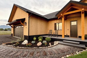 Een houten huis met twee verdiepingen, een garage, een veranda met stoelen en een aangelegde voortuin met rotsen en planten.