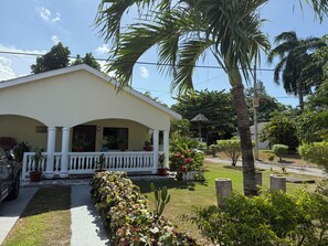 A two-story house with a porch, surrounded by a garden with various plants and a palm tree. - Lydia’s Loft- A Cozy Private 2-Bedroom Guest Suite Located near Drax Hall (Saint Ann's Bay)