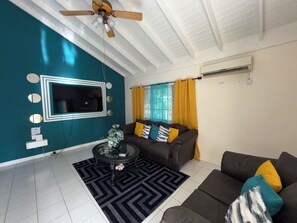 A living room with a ceiling fan, a flat-screen TV mounted on a teal wall, a brown sofa with yellow cushions, and a patterned rug. - Lydia’s Loft- A Cozy Private 2-Bedroom Guest Suite Located near Drax Hall (Saint Ann's Bay)