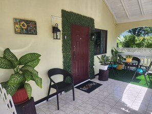 A covered outdoor area with a red door, a black chair, a potted plant, a welcome mat, and a small table with chairs. - Lydia’s Loft- A Cozy Private 2-Bedroom Guest Suite Located near Drax Hall (Saint Ann's Bay)