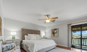 Ein Schlafzimmer mit einem großen Bett, einem Deckenventilator, einem Nachttisch mit Lampe, einem Gemälde und Seeblick durch ein Fenster.