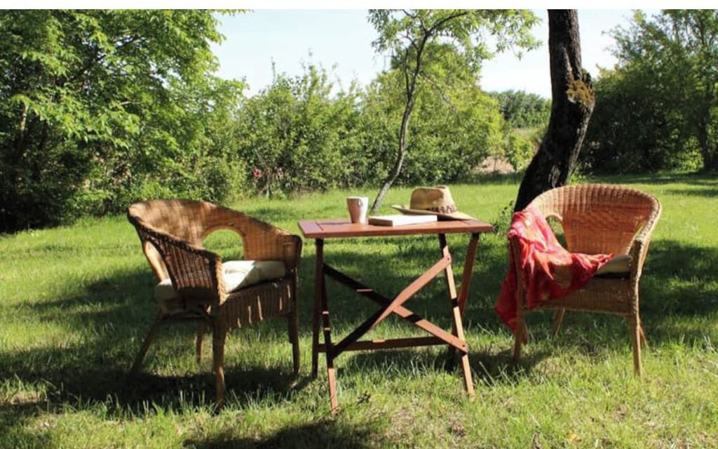 An outdoor seating area with wicker chairs and a wooden table, surrounded by greenery.