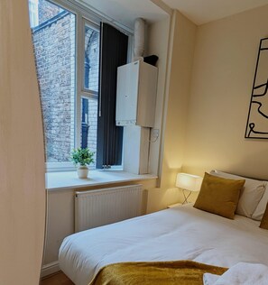 A hotel room with a bed, a window with a view of a brick building, a radiator, a nightstand with a lamp, and a wall-mounted air conditioning unit. - City Centre 2 Bedroom Apartment Liverpool (Liverpool)