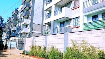 Um edifício residencial com vários andares, varandas, entrada com portão e uma estrada de terra em frente.