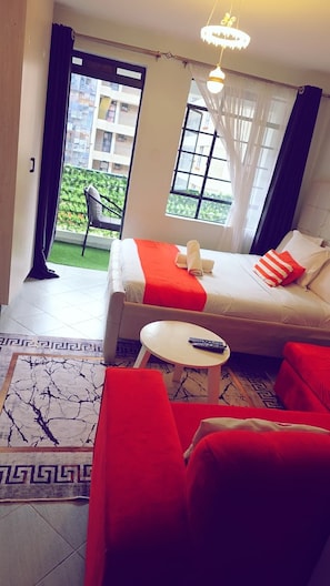 A hotel room with a bed, a small table, a chair, and a view of a balcony with a chair outside. - Amaziah Apartments Muthiga (Rungiri)